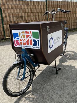 Lastenrad auf einem Parkplatz. Vorne auf der Kiste ist das Logo des Global Goals Radwegs zu sehen, links ist das Welthaus Logo abgebildet.
